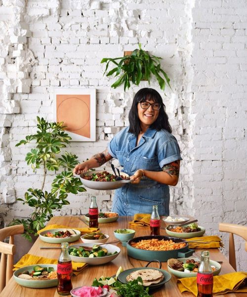 Adult woman with glasses holding a pan of food in a dining room with a set table with bowls of food, Coca-Cola bottles, and plants.