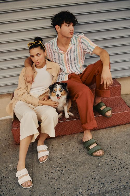 Two people and a dog sit on stairs in an urban setting. The individuals are wearing casual clothing and sandals in a medium outdoor shot.
