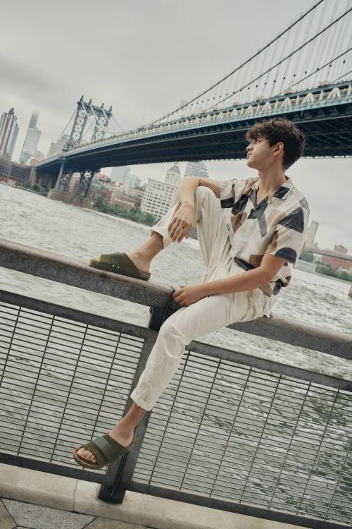 Man sits on metal railing with city bridge and skyline backdrop, wearing casual shirt, trousers, and green sandals, gazing upwards.