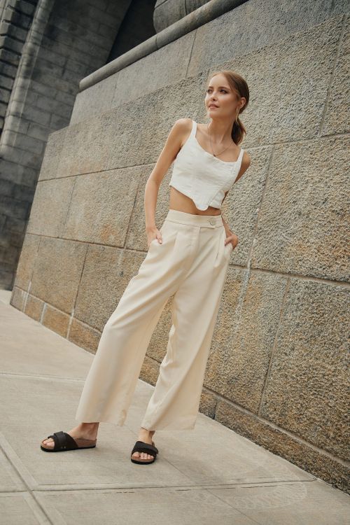 Blonde woman poses against a concrete wall wearing a white crop top and beige linen pants with slide sandals in an urban setting.