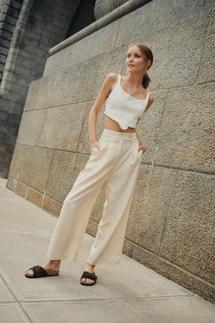 Blonde woman poses against a concrete wall wearing a white crop top and beige linen pants with slide sandals in an urban setting.