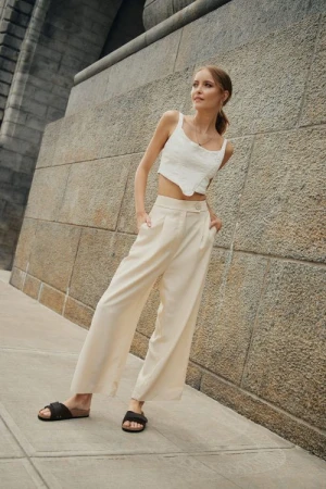 Blonde woman poses against a concrete wall wearing a white crop top and beige linen pants with slide sandals in an urban setting.