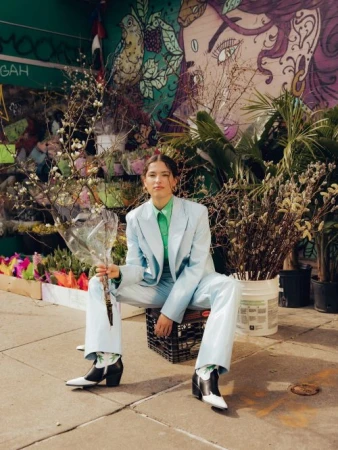 A woman in a blue suit poses with flowers in an urban outdoor setting with a mural backdrop, showcasing fashion and street style.