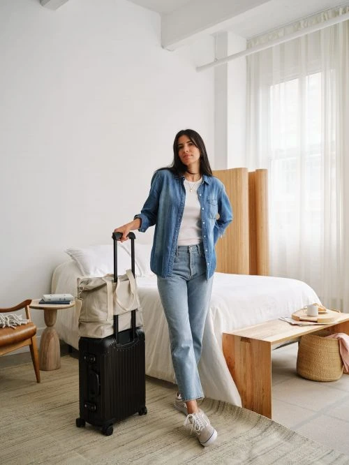 Woman in denim shirt and light blue jeans posing with black rolling luggage and a duffel bag in a bedroom setting.