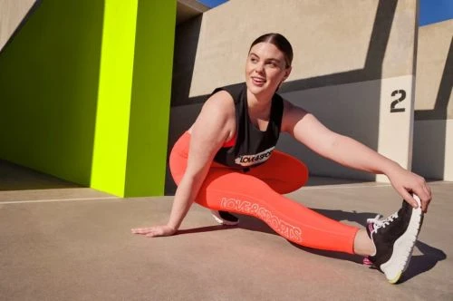 Woman in vibrant orange leggings and black athletic top stretching outdoors against a concrete wall