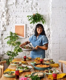 Woman in denim jumpsuit holding a large serving dish at a wooden dining table laden with food and Coca-Cola bottles.