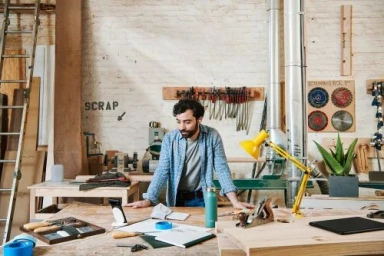 Bearded man in a blue plaid shirt leaning over a workbench in a workshop styled by Courtney Raniszewski