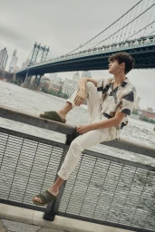 Young man in patterned short-sleeve shirt and light trousers sitting on a metal railing by a river with a suspension bridge