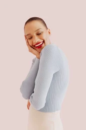 Close-up portrait of a smiling woman with short hair wearing a light blue ribbed sweater against a solid pink backdrop