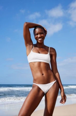 Black woman with a shaved head wearing a white bikini posing on a sandy beach against an ocean and blue sky.