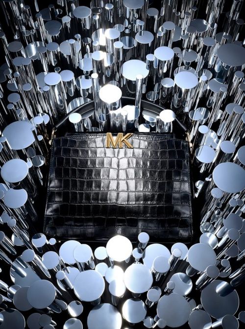 Close-up of a black, textured Michael Kors handbag with gold logo against a background of mirrored cylindrical shapes.