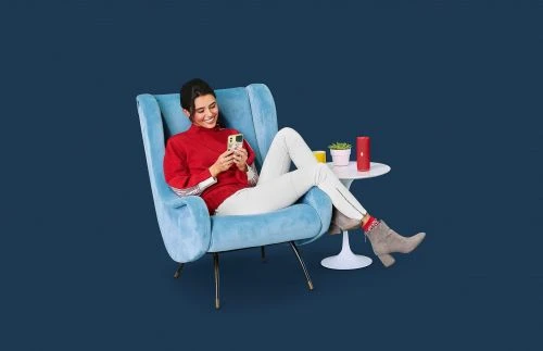 A woman sits in a blue wingback chair, smiling and looking at her cell phone in an indoor studio setting with a dark blue background.