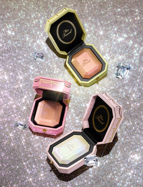 Three Too Faced powder compacts in yellow, pink, and white shimmer on a sparkling glittery background with diamond prop accents.