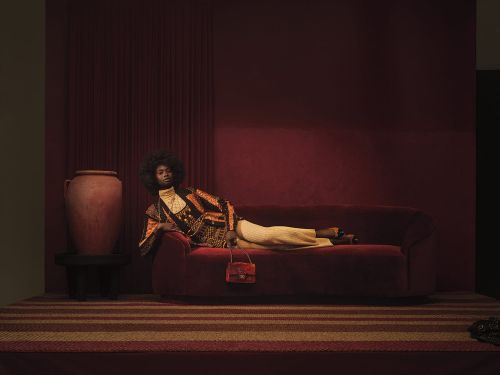 A Black fashion model lounges on a maroon velvet sofa with a patterned blazer, cream pants, and a mini purse in an indoor studio.