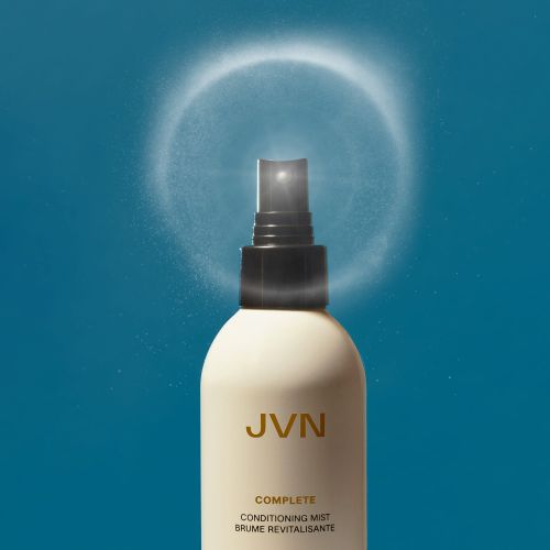 Close-up of JVN Complete Conditioning Mist spray bottle on blue background with visible mist spraying from nozzle in circular pattern.