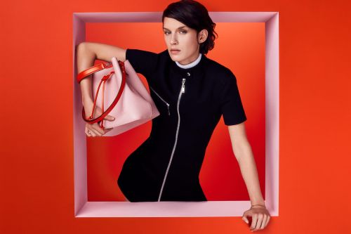 Fashion shot of a woman in a black dress, holding a light pink bucket bag with orange handles, posing in a pink-trimmed frame.