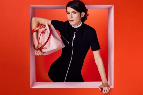 Fashion shot of a woman in a black dress, holding a light pink bucket bag with orange handles, posing in a pink-trimmed frame.