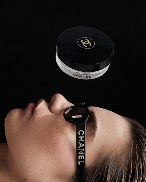 Beauty shot of a woman lying down wearing branded Chanel sunglasses, with Chanel Les Beiges powder floating above, against black.