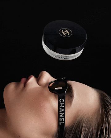 Beauty shot of a woman lying down wearing branded Chanel sunglasses, with Chanel Les Beiges powder floating above, against black.