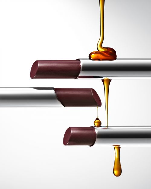 Studio shot of three lipsticks arranged horizontally, with honey dripping down the side of the tubes on a clean white background.