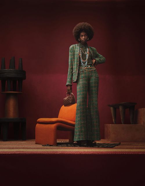 A stylish Black woman with an afro poses in a green tweed suit in a studio setting with a retro orange chair and interesting furniture.