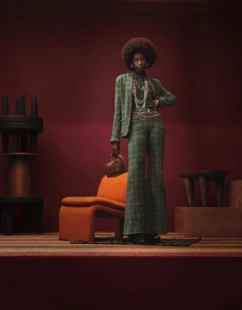 A stylish Black woman with an afro poses in a green tweed suit in a studio setting with a retro orange chair and interesting furniture.