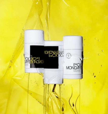 Three white deodorant sticks with black and gold labels, arranged against a bright yellow background with translucent plastic sheets.
