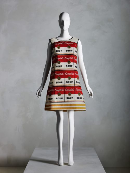 Mannequin displays dress featuring Campbell's Soup cans pattern against a mottled gray background in a studio setting.