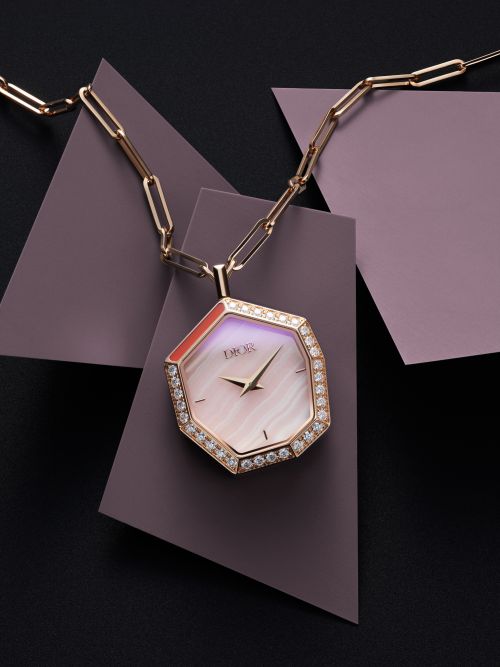 Elegant Dior watch necklace with a diamond bezel on a gold chain against a geometric mauve and black background in a studio setting.