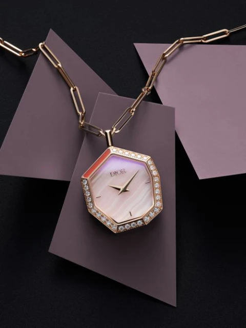 Elegant Dior watch necklace with a diamond bezel on a gold chain against a geometric mauve and black background in a studio setting.