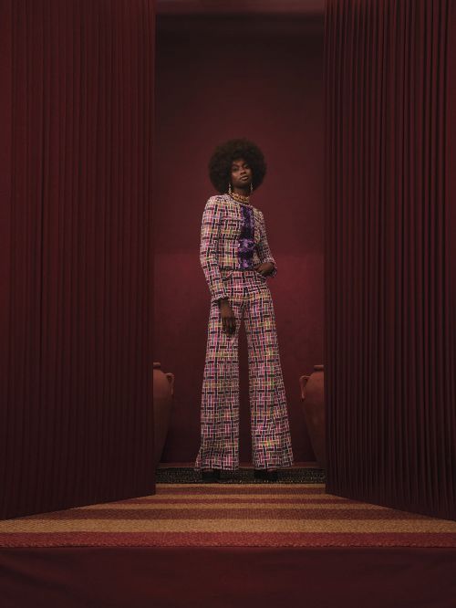 A stylish Black woman in a patterned suit poses in front of a red curtain, flanked by pottery, against a monochromatic backdrop.