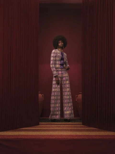 A stylish Black woman in a patterned suit poses in front of a red curtain, flanked by pottery, against a monochromatic backdrop.