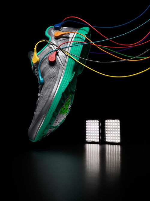 Studio shot of a gray and green running shoe adorned with colored wires and electrical connectors, against a black backdrop with LED lighting.