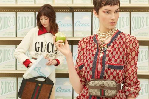 Two fashion models in a grocery setting wear Gucci attire and accessories, with a backdrop of stacked laundry detergent boxes.