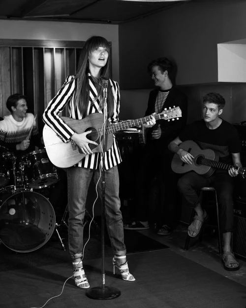 Black and white shot of a woman with long hair playing a guitar and singing into a microphone, backed by three other musicians.