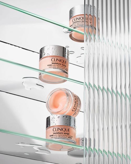 Three jars of Clinique Moisture Surge cream on glass shelves, with water droplets, against a white background and textured glass panel.