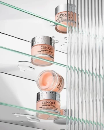 Three jars of Clinique Moisture Surge cream on glass shelves, with water droplets, against a white background and textured glass panel.