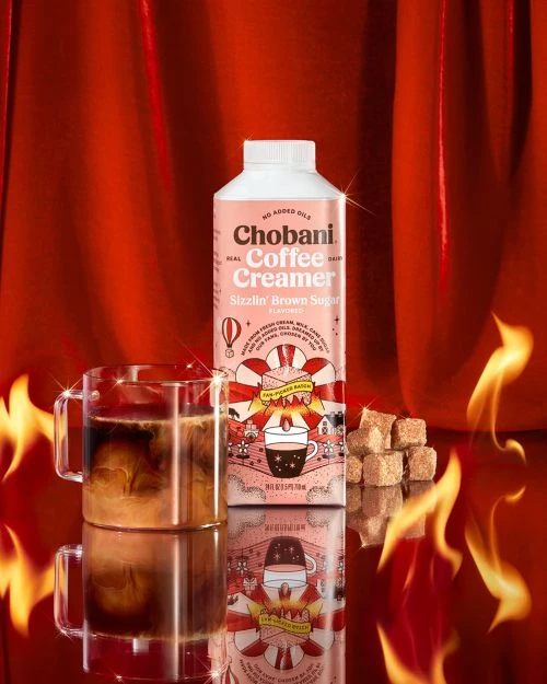 Chobani Coffee Creamer 'Sizzlin' Brown Sugar' on a reflective surface with sugar cubes and two stacked glass coffee mugs.