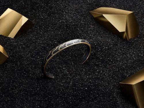 An ornate metal bracelet with a geometric pattern sits among gold geometric shapes on a textured black surface in a studio setting.