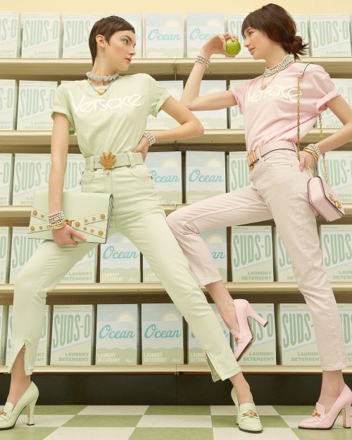 Two women posing in front of rows of detergent boxes, wearing matching designer shirts and pants with an apple and purse.