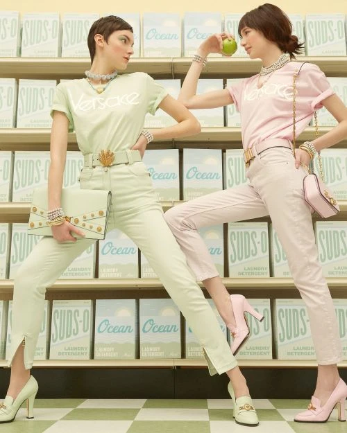 Two women posing in front of rows of detergent boxes, wearing matching designer shirts and pants with an apple and purse.