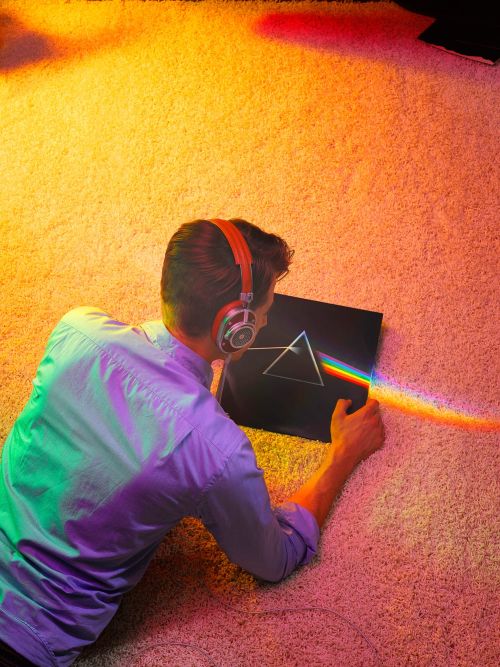 Man lying on carpet with orange headphones, holding the cover of Pink Floyd's 'Dark Side of the Moon' album, bathed in colored light.