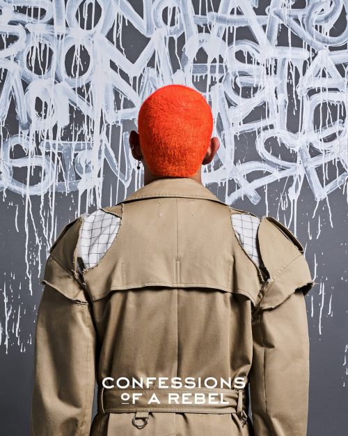 An adult from the back, sporting an orange haircut and beige trench coat, stands against a gray wall featuring abstract graffiti art.