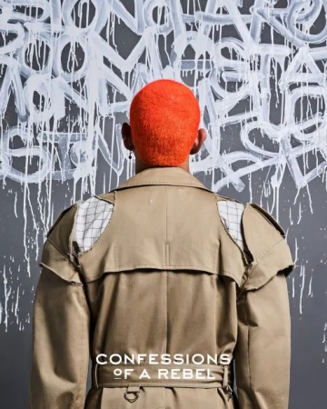 An adult from the back, sporting an orange haircut and beige trench coat, stands against a gray wall featuring abstract graffiti art.