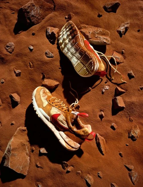 Two athletic shoes, one upside down, resting on red-brown sand and rocks, suggesting a desert or Martian landscape in a close-up shot.