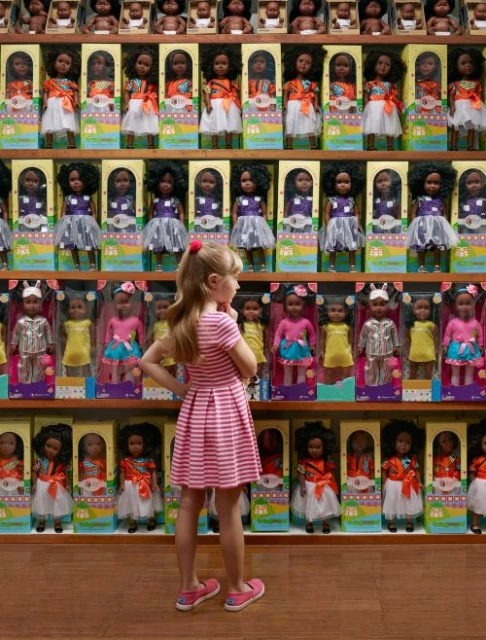 Young girl in a pink and white striped dress stands viewing a display of fashion dolls in boxes arranged on shelves in a store.