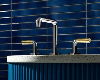 Chrome faucet with textured gold handles mounted on a stone countertop against a blue tiled wall.