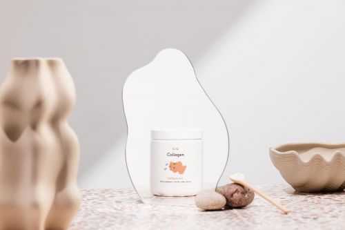 Still life shot of collagen jar, stone, shell, and wooden spoon against a white and neutral-tone background with subtle shadows.