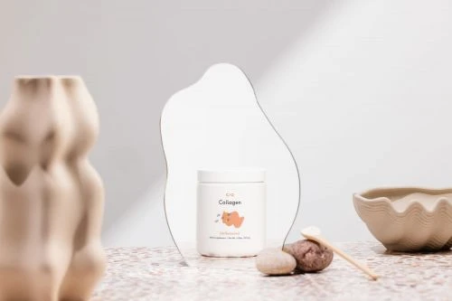 Still life shot of collagen jar, stone, shell, and wooden spoon against a white and neutral-tone background with subtle shadows.