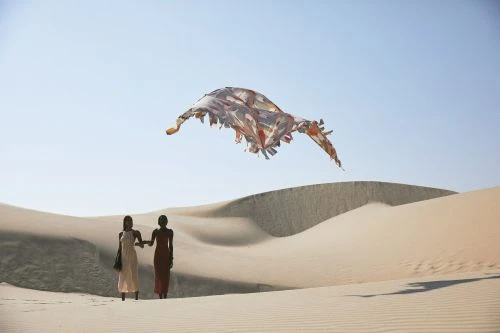 Two women in dresses holding hands walk through a sandy desert landscape beneath a large, colorful textile floating in the sky.